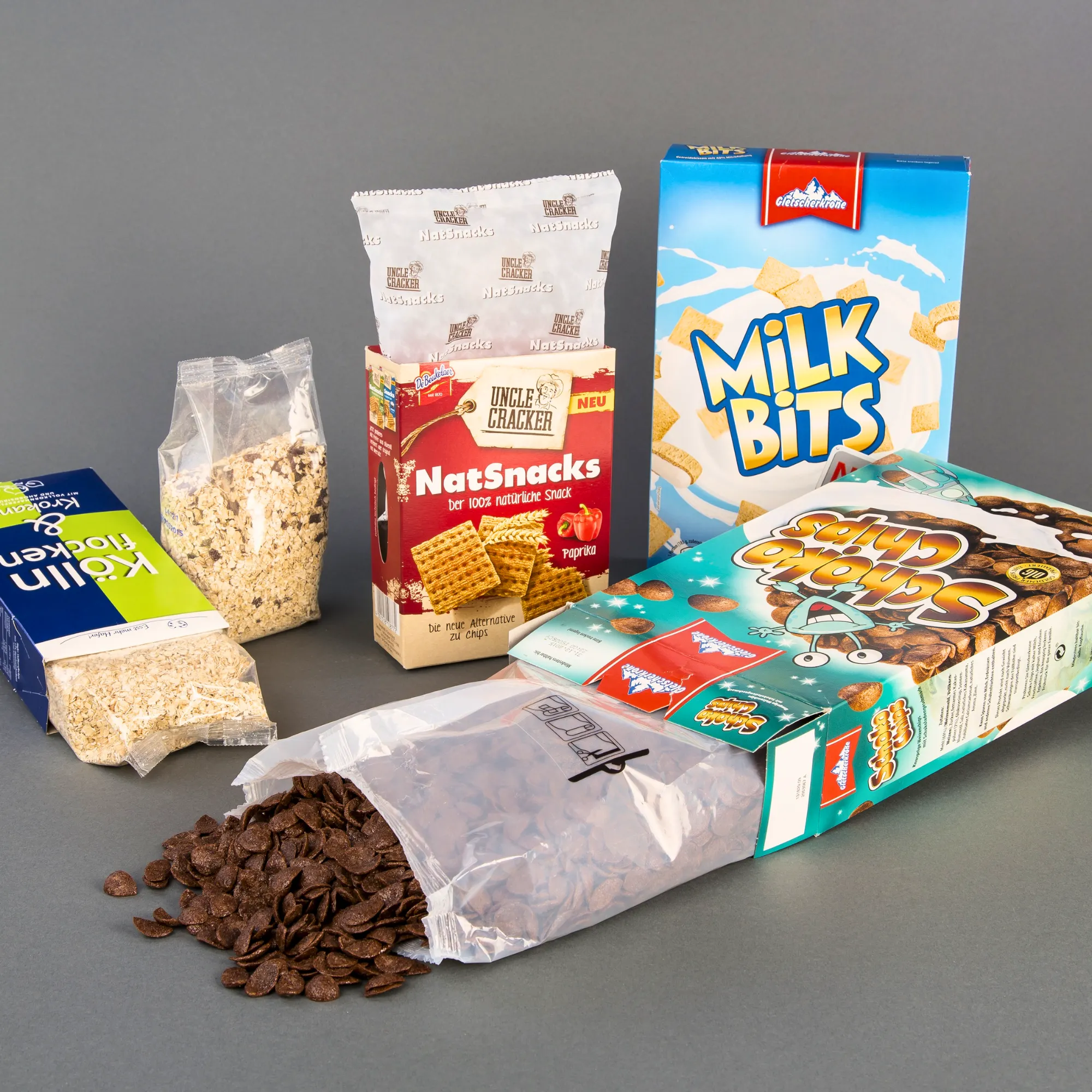 Various cereal and snack packages arranged on a gray background, including Milk Bits and assorted snacks.