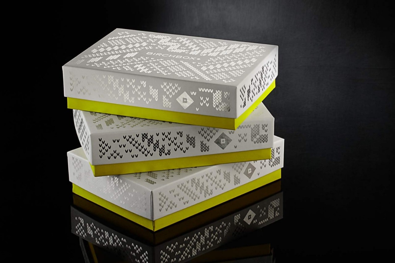Three stacked Birchbox gift boxes with a decorative pattern, featuring a white exterior and vibrant yellow base, set against a dark background.