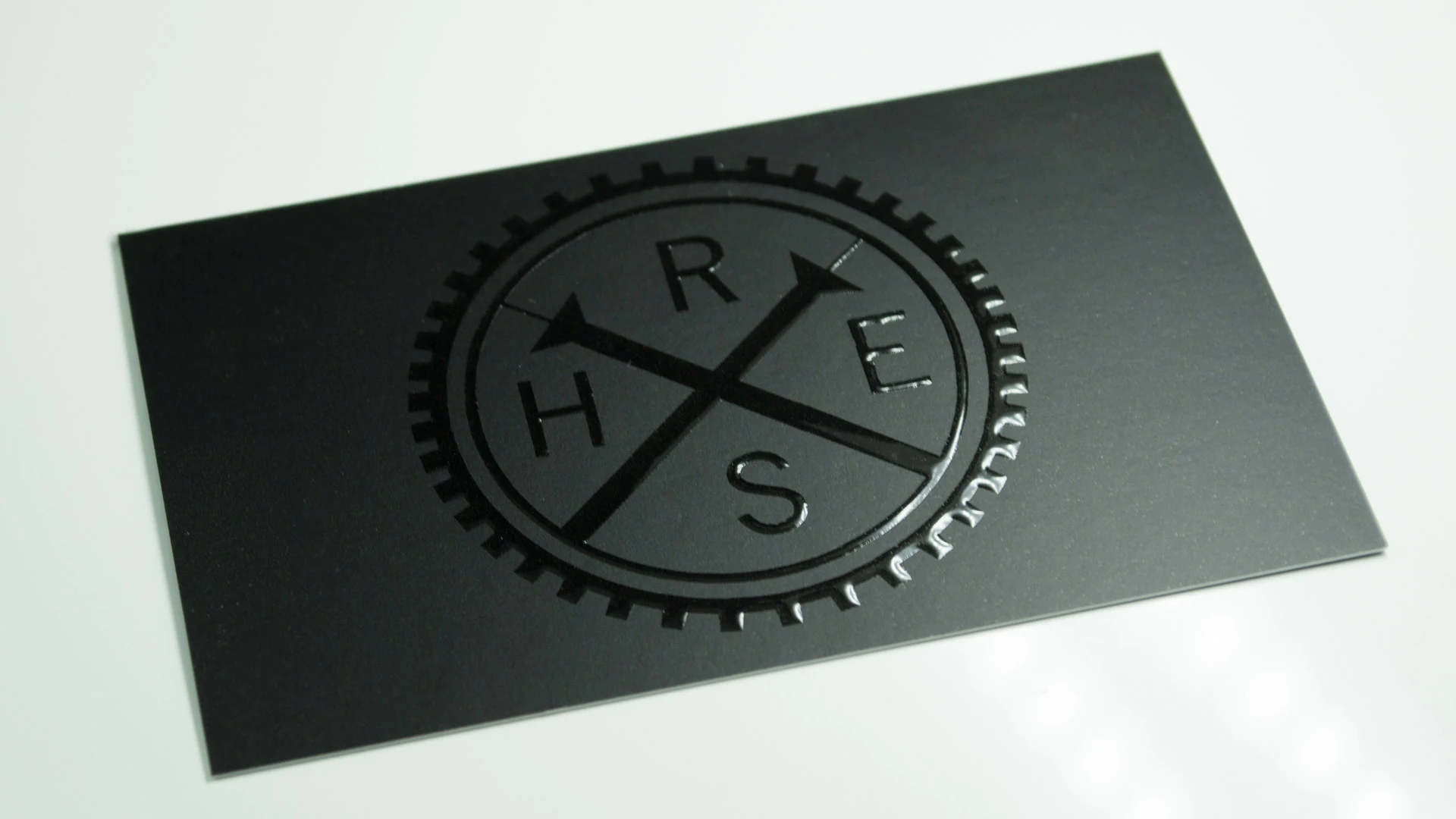 A close-up of a black card featuring a circular logo with the letters "R", "H", "S", and "E" arranged in a cross formation, surrounded by a gear-like border.