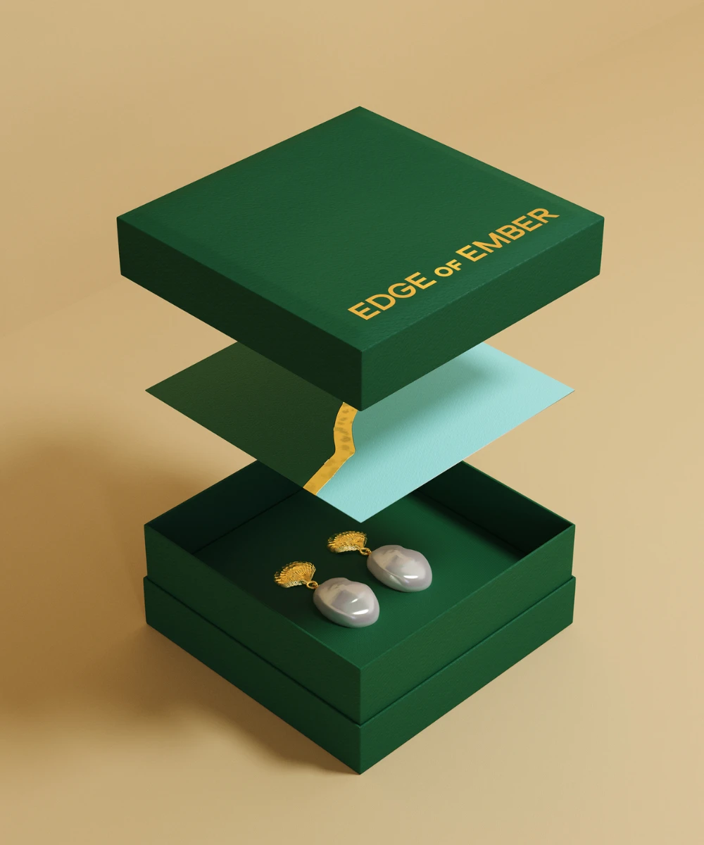 A luxurious green jewelry box from EDGE of EMBER is displayed with its lid slightly opened, revealing a pair of elegant earrings resting inside on a light tan background.