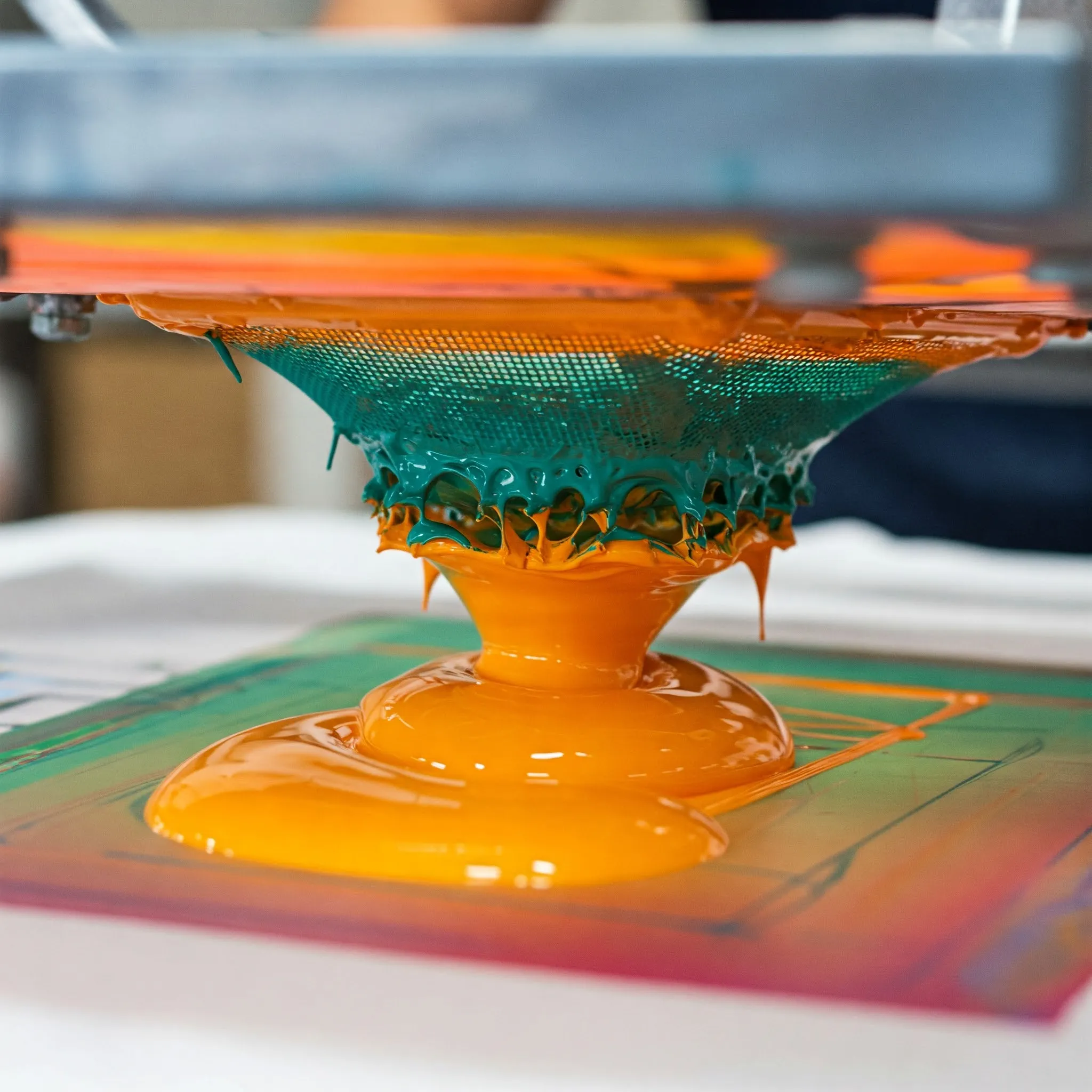 Close-up view of vibrant screen printing inks in orange and teal, with a mesh screen capturing dripping paint.